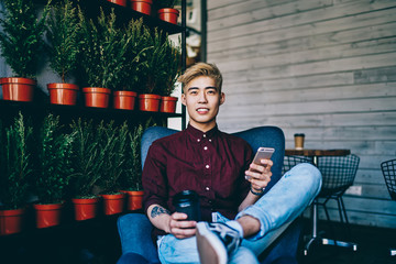Fototapeta na wymiar Positive chinese hipster blogger holding coffee to go in hand while resting in comfortable chair in modern cafe with loft interior, cheerful asian young man using smartphone device for communication