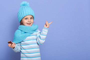 Little girl standing isolated over blue wall points at space aside with her index finger and looking at camera, female child holds chocolate candy in hand, wearing, cap with pom pom, scarf and shirt.