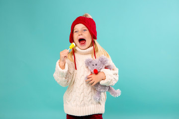 little blonde girl in a red knitted hat and sweater holding a soft toy mouse and a yellow Lollipop. isolate on blue background, space for text