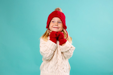 little blonde girl in a red knitted hat, mittens and a white sweater smiles on a blue background. winter clothes, space for text