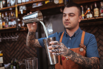 Attractive tattooed bartender mixing ingredients in steel shaker, making cocktail for a customer