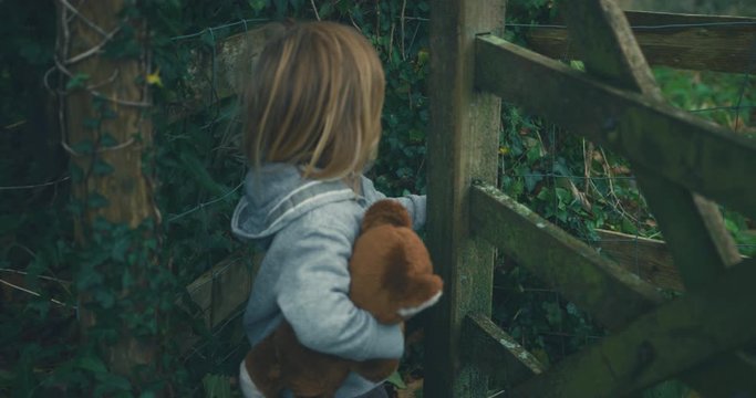 Little toddler with teddy bear opening a gate in the woods