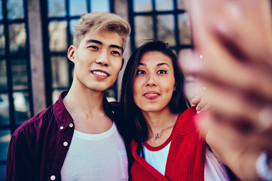 Cheerful chinese hipster girl with funny face making grimace to take selfie together with best friend on street.Positive asian couple in love standing outdoors and spending time together