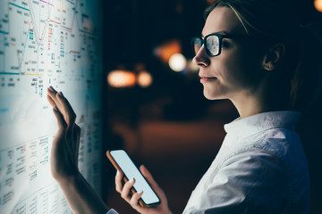 Digital monitor with public transport subway map. Female standing at big display with smartphone in hand. Young woman touching with finger screen while using train schedule application on mobile phone