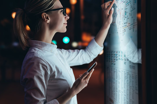 Interactive Kiosk With Public Transport Subway Map.Female Standing At Big Display With Smartphone In Hand.Young Woman Touching With Finger Screen While Using Train Schedule Application On Mobile Phone