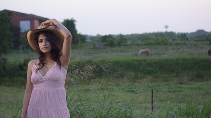 Beautiful woman sitting on a fence staring down at the ground with her hands on her hat