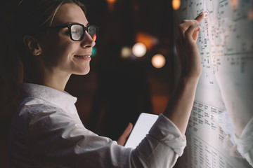 Interactive kiosk with public transport subway map.Female standing at big display with smartphone in hand.Young woman touching with finger screen while using train schedule application on mobile phone