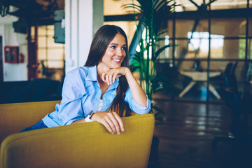 Positive smiling female student 20s sitting indoors and enjoying free time on leisure, caucasian successful woman casual dressed smiling and looking away while waiting friend in college campus