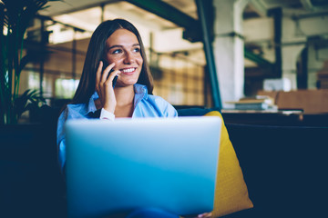 Happy female freelancer enjoying distance job indoors during positive cellular conversation, successful hipster girl talking with friend via smartphone device while holding laptop computer on legs
