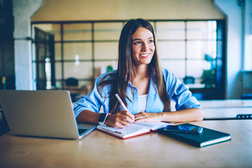 Smiling successful woman with netbook enjoying planning process while sitting at desktop with modern laptop device indoors, young positive female student thinking about ideas for school project