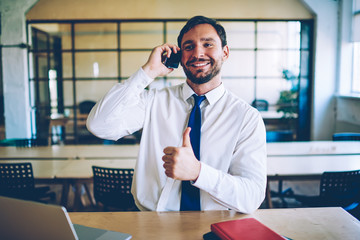 Happy male entrepreneur communicate with colleague via cellular phone during working time in office showing super on hand, positive man in formal wear communicate via smartphone gadget indoors