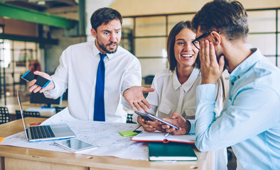 Positive male and female partners laughing and enjoying live communication while angry colleague near talking to look in smartphone gadget during break, concept of collaboration and cooperation