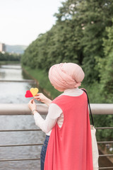 romantic portrait of young European Muslim woman with hijab have fun at beautiful sunny day. Image of pretty woman playing with two heart shaped lollipops,standing on the bridge and looking into river