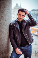 One handsome young man in urban setting in modern city, standing, wearing black leather jacket and jeans, looking at camera