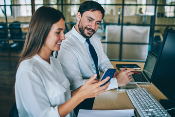 Positive male and female employees watching funny video online via smartphone gadget sitting at desktop with computers, successful colleagues enjoying communication togetherness reading news
