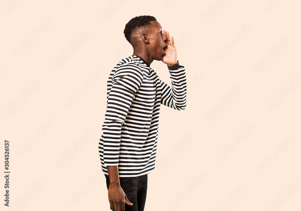 Wall mural young african american black man profile view, looking happy and excited, shouting and calling to copy space on the side against beige wall