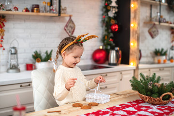 cute baby in light sweater and headband with reindeer horns sitting at kitchen table smiling opens gift box