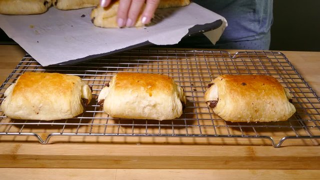 Closeup shot of a woman&rsquo;s hand transferring pain au chocolat pastries / chocolate croissants, on a baking tray, hot from the oven, onto a cooling grid.