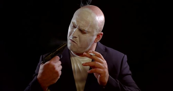 portrait of a man with a face covered with white makeup on a black background. he smokes a cigarette, looks discontentedly at the gun in his hand, blows smoke into the barrel, inhales
