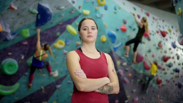 Waist-up arc portrait shot of confident tattooed Caucasian female climbing coach posing for camera with folded arms in indoor climbing gym, and three trainees bouldering in background