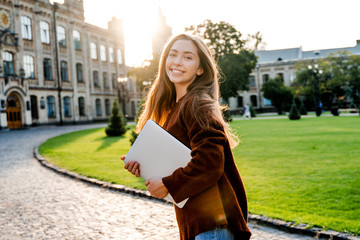 Charming young woman student in move walking in university garden with laptop computer, going to her classroom, beautiful sunset background, positive emotions, lifestyle people