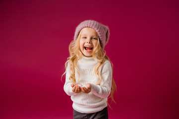 little blonde girl in a knitted hat and sweater catches the snow and laughs. winter clothes. isolate on red background, space for text