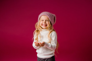 little blonde girl in a knitted hat and sweater catches the snow and laughs. winter clothes. isolate on red background, space for text