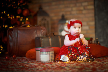 little girl in red dress against background of Christmas tree holds Christmas garland in her hands. baby 6 month old celebrates Christmas.