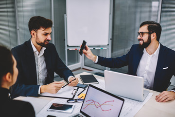 Cheerful bearded financial manager showing smartphone with funny business news to colleague during brainstorming in office.Successful boss watching video on telephone collaborating with employees