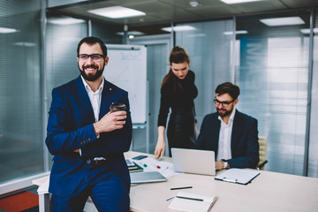 Handsome successful executive manager enjoying delicious coffee break during sitting on table while intelligent colleagues on background creating presentation using application on laptop computer