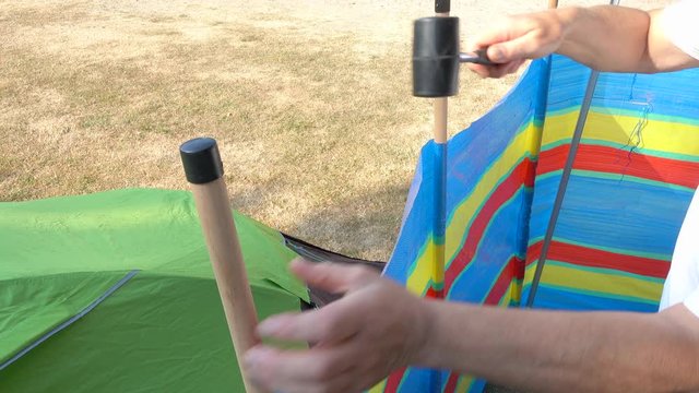 POV close shot of a man&rsquo;s hands securing wooden windbreak poles next to a small tent on a campsite, by hammering the tops with a rubber mallet.