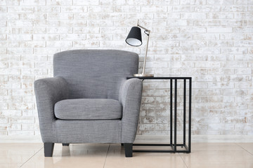 Armchair with table and lamp near brick wall in room