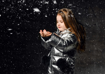 Little girl kid in a winter silver jacket down jacket stands under the snow and catches snowflakes...