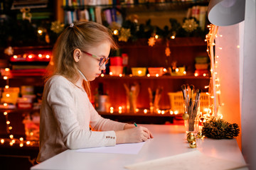 Adorable litle girl is writting a letter to Santa Klaus sitting on the table