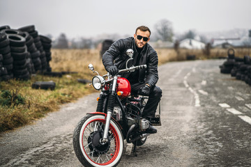 Obraz na płótnie Canvas Bearded man in sunglasses and leather jacket looking at the camera while sitting on a motorcycle on the road. Behind him is a row of tires