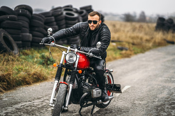 Bearded man in sunglasses and leather jacket looking at the camera while sitting on a motorcycle on the road. Behind him is a row of tires