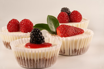 Front view and close-up of homemade cheesecakes and homemade strawberry sauce accompanied by strawberries, blackberries, raspberries and green leaves on white background.