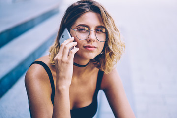 Close up portrait of attractive hipster girl in trendy sunglasses looking at camera during international smartphone conversation, charming woman calling to operator during free time outdoors