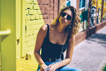 Portrait of confident female meloman in sunglasses looking at camera while listening music from...