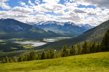 A view of the green grassy hills overlooking Banff mountain range with the valleys below covered trees and wetlands on a partially sunny day in Banff, Canada.