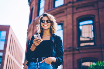 Half length portrait of cheerful hipster girl looking at camera while using smartphone for creating travel publication, smiling woman in sunglasses enjoying spring day and good internet connection