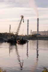 Industrial landscape with cranes and pipes. The plant works near the river. Crane on a barge loads cargo. Industrial area. Cityscape Scenery.