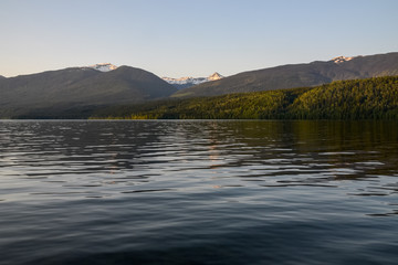 A warm sunset glow tints the clear sky, highlighting the snow capped mountain peaks and dense pine forest beyond the reflective lake, skewed its rippled surface.