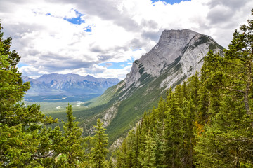 Peering through the pine trees on a mountain hiking trail, the rocky Mount Rundle stands tall and proud amongst the forest with a lake and mountain range in the background on a sunny and cloud day.