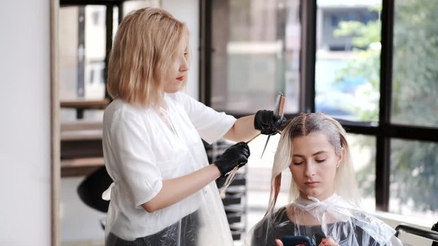 A hairdresser has a short blond hair. She wears the black gloves. The girls are in the beauty studio.
