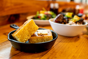 Cornbread in a cast iron pan, with Pineapple Grilled Chicken Garnished with Green Onion, with side of Wild Rice and Fried Plantains in a bowl to the side and other food blurred in the background