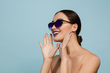 Portrait of beautiful woman with bright make-up and sunglasses on blue studio background. Stylish, fashionable make and hairstyle. Colors of summer. Beauty, fashion and ad concept. Calling somebody.