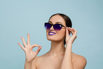 Fototapeta premium Portrait of beautiful woman with bright make-up and sunglasses on blue studio background. Stylish, fashionable make and hairstyle. Colors of summer. Beauty, fashion and ad concept. Shows sign of nice.