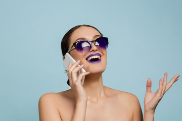 Portrait of beautiful woman with bright make-up and sunglasses on blue studio background. Stylish and fashionable make and hairstyle. Colors of summer. Beauty, fashion and ad concept. Talking on phone