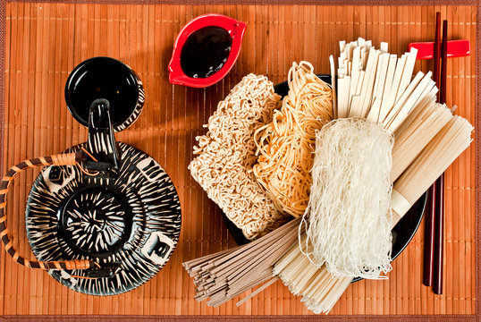 Different bundles of uncooked asian noodles (udon, soba, ramen, rice and glass noodles) in a bowl with chopsticks, soy sauce and teapot on a bamboo background. Top view.